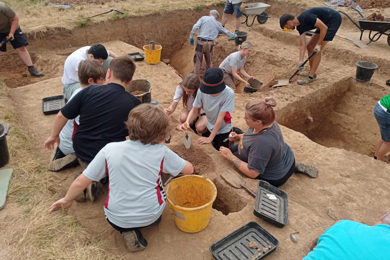 A group of children and adults gather around various excavated holes in the ground with buckets and shovels.