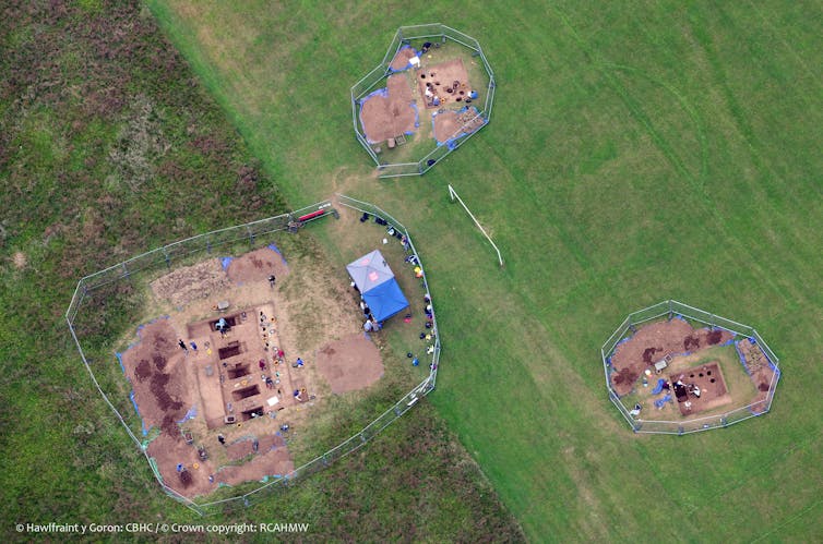 An aerial shot of a green field with three separate archaeological excavations taking place. There are precise holes in the ground and a blue tent set up.