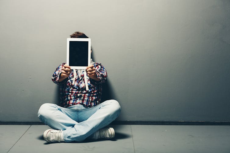 Young man in jeans and trainers sitting cross-legged on the ground in front of a grey wall holding up a blank tablet in front of his face