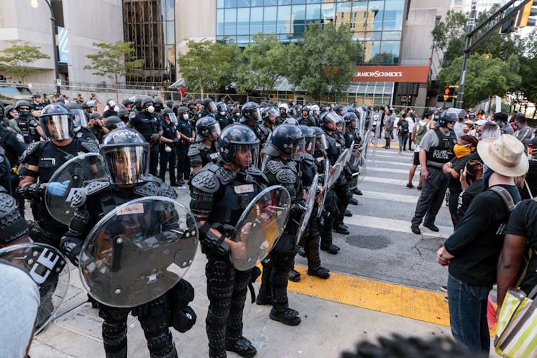 Rows of armed police in riot gear with face and body shields at an outdoor protest