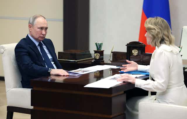 Russian president Vladkmir Putin sits at a desk opposite Russia's children's commissioner Maria Lvova-Belova.