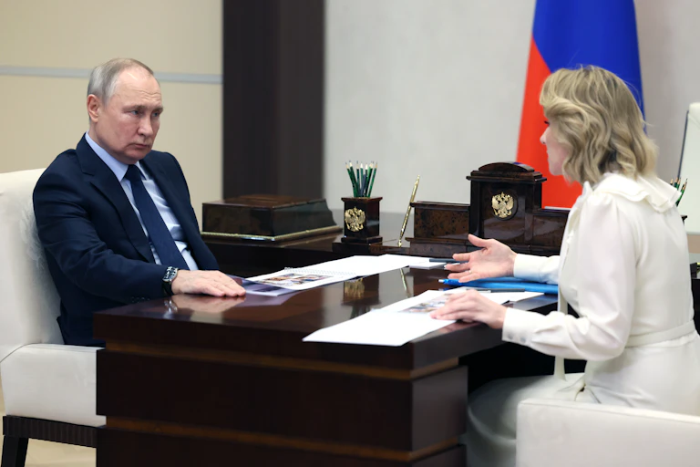 Russian president Vladkmir Putin sits at a desk opposite Russia's children's commissioner Maria Lvova-Belova.