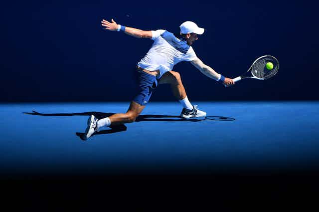 Tennis player Novak Djokovic playing in white and navy, reaching for a backhand on a blue court.