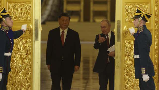 Two men gesture as they walk through golden doors opened by two men in uniform