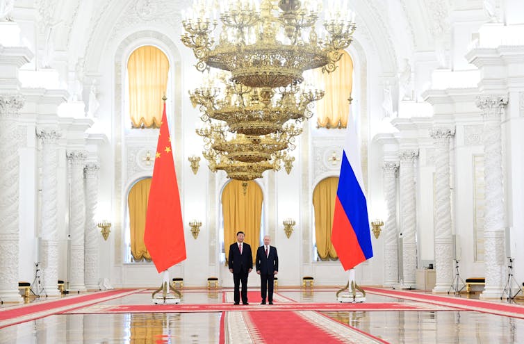 Two men are seen in the background flanked by giant China and Russian flags. Chandeliers hang overhead.