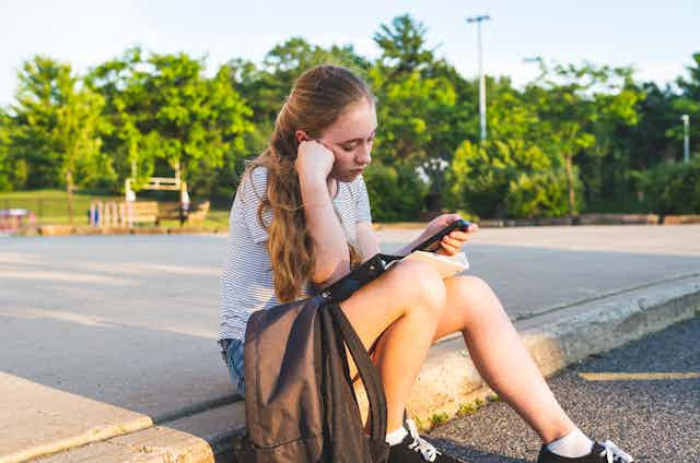Girl sitting on pavement looking sadly at phone