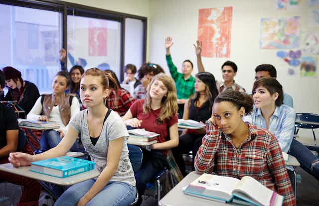 High school students in a classroom are seated at their desks. Some of them have raised hands.