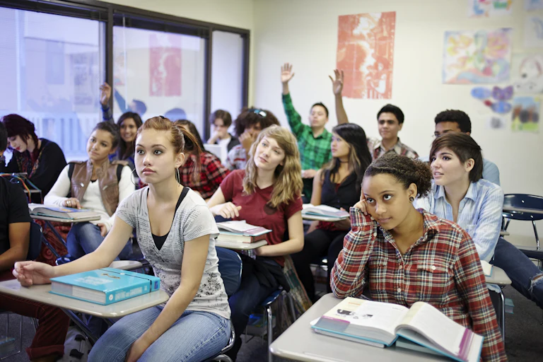 High school students in a classroom are seated at their desks. Some of them have raised hands.