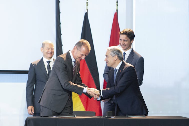 Two men shake hands while two other men look on. A Canadian flag and a German flag stand in the background.