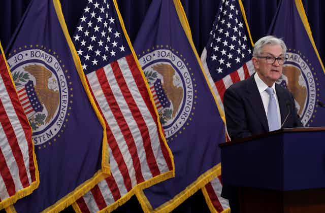 A man stands by a lectern in front of several US and Federal Reserve flags.