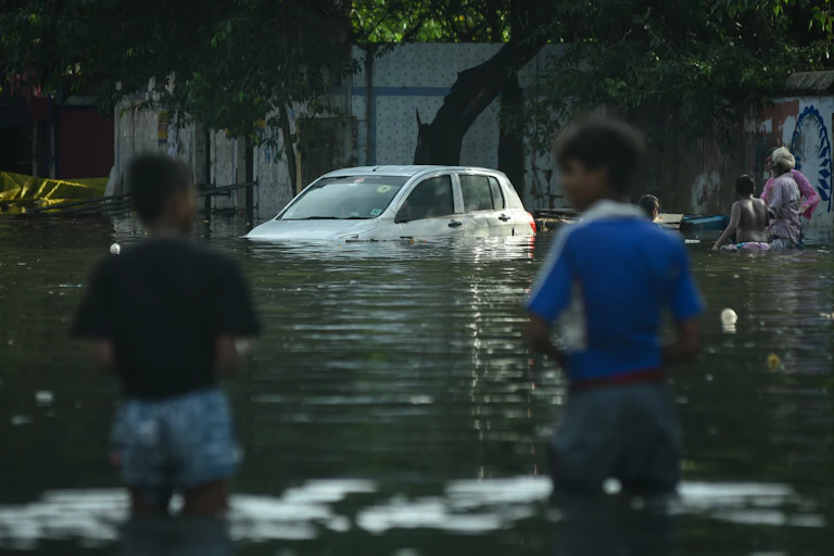 Two children stand in knee-deep water with a submerged car in the distance.