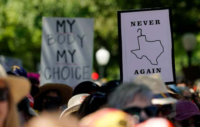 Protesters hold up signs, one depicting the map of Texas as a coat hanger that says Never Again, another says My Body My Choice.
