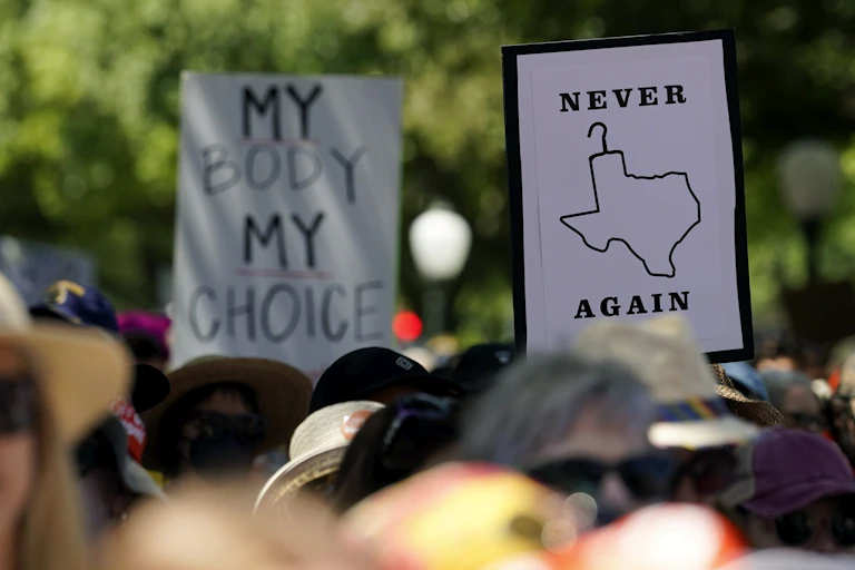 Protesters hold up signs, one depicting the map of Texas as a coat hanger that says Never Again, another says My Body My Choice.