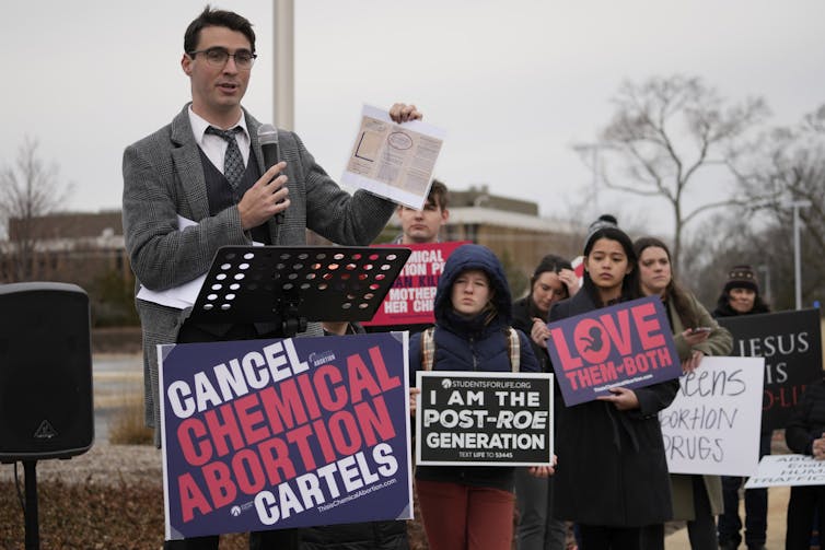 A man speaks into a microphone behind a sign that reads Cancel Chemical Abortion Cartels.