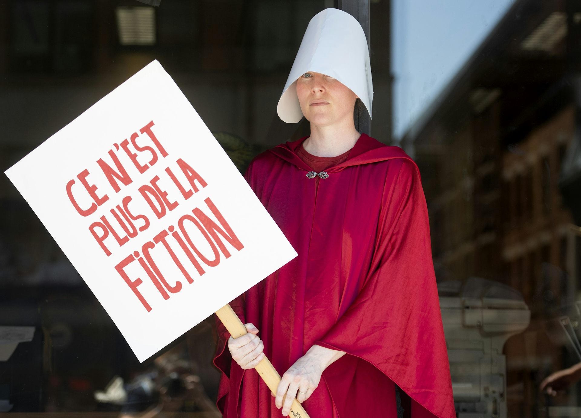 A woman dressed in red robes and a white bonnet holds a French sign that read It's No Longer Fiction.