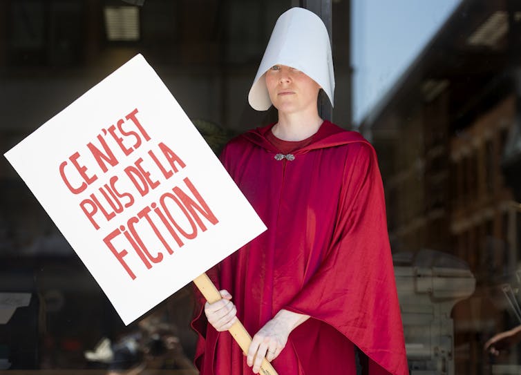 A woman dressed in red robes and a white bonnet holds a French sign that read It's No Longer Fiction.