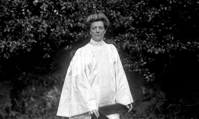 A black and white photo of a woman in a white robe standing in a garden.