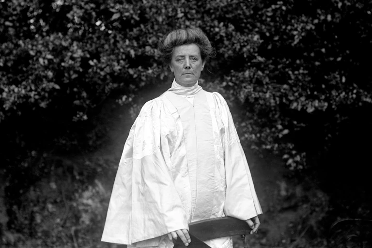 A black and white photo of a woman in a white robe standing in a garden.