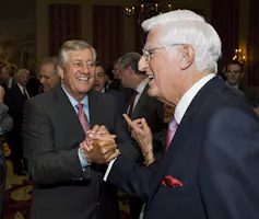 A white-haired man with glasses shakes hands with another smiling man.