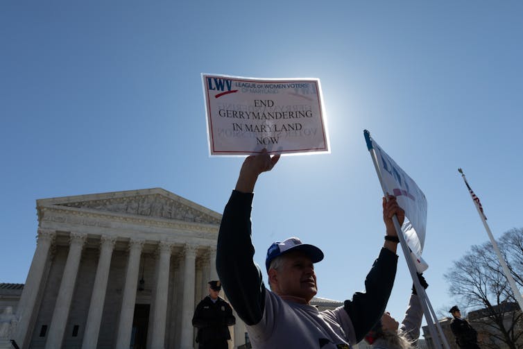 A man in front of a white stone building with columns, wearing a baseball cap holding a sign 'End gerrymandering in Maryland.'