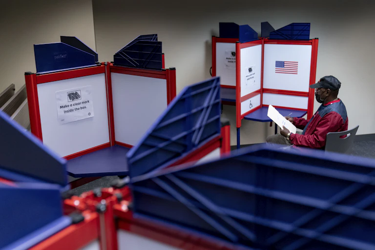 A manin a red and blue jacket holding a rectangular ballot at a table.
