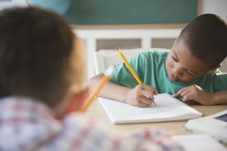 A boy writes on a booklet while seated at a desk.