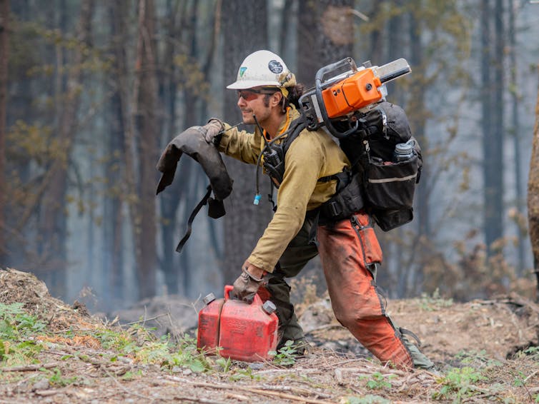 A firefighter in the woods loaded with gear, including chain saw, fuel canister and full backpack.