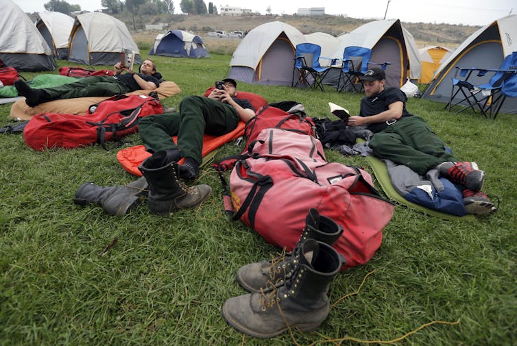 Trois pompiers se prélassent sur des matelas pneumatiques tout en lisant.  Les tentes sont derrière eux et les bottes sont au premier plan.