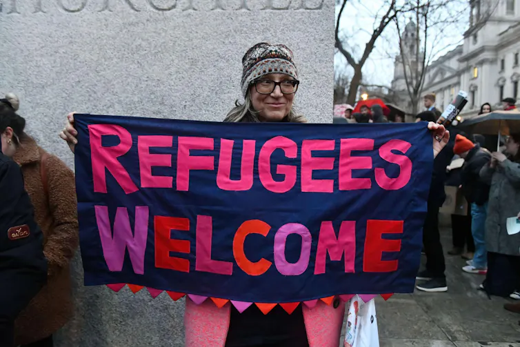 A protester holds a banner reading 'Refugees Welcome' in pink and purple letters.