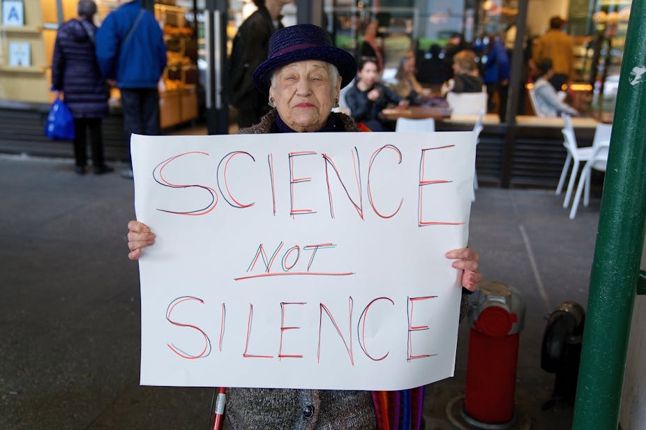 An elderly lady wearing a purple hat holds up a sign which says 'science not silence'.