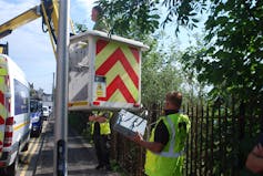 A man in a high vis jacket with a clipboard stands in front of a cherry picker. Another man stands in the cab next to a street light.