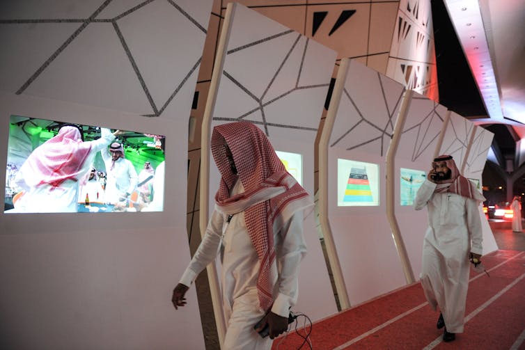 Saudi men walk down hallway past an exhibition with digital screens