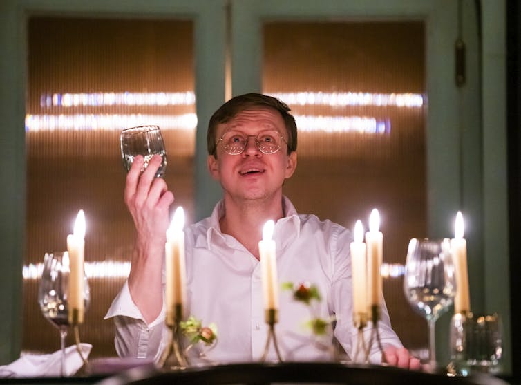 A man sits in a white shirt, lit by candles as he raises his glass as if in a toast.