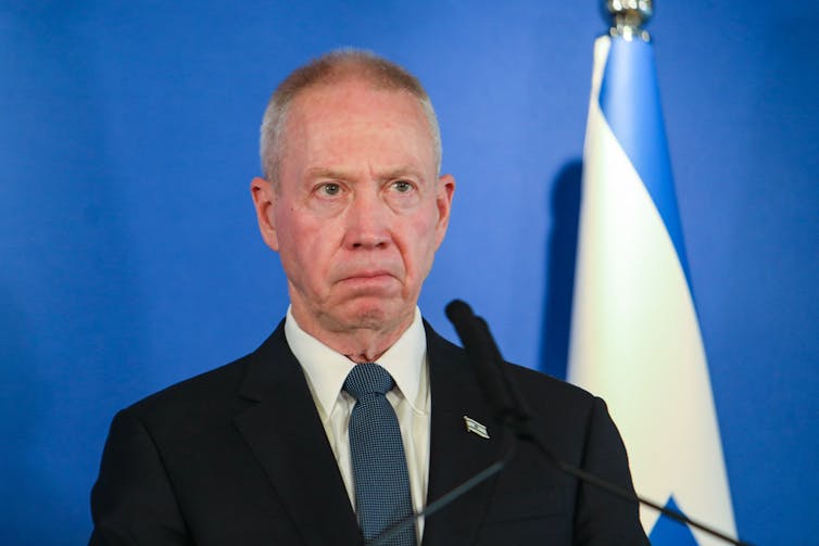 A man with gray hair, wearing a suit jacket and tie, standing in front of a blue wall and blue and white flag.