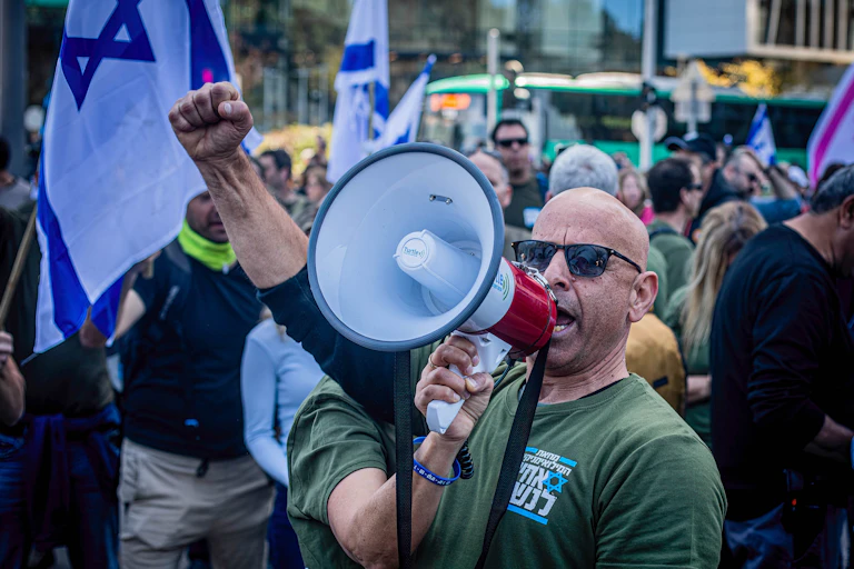 A bald man in green shirt and sunglasses speaks into a bullhorn and raises his arm in a fist.