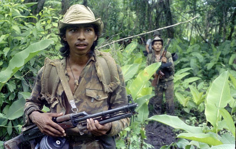 Two heavily armed soldiers walk through a jungle.
