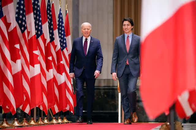 An older man walks next to a younger man with dark hair with U.S. and Canadian flags on either side of them. 
