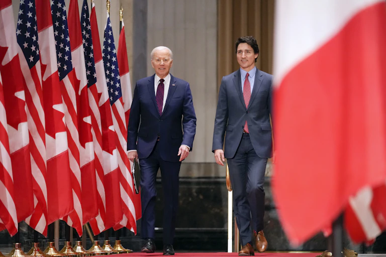 An older man walks next to a younger man with dark hair with U.S. and Canadian flags on either side of them.