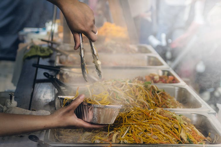 A row of fried noodle dishes with a person filling up a foil container in the foreground