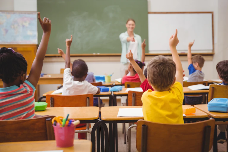 A school room with young pupils raising their hands.