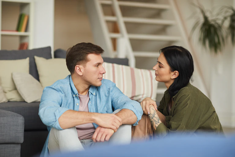 A young man and a young woman in their 20s or 30s sit on the floor in front of their couch talking honestly to each other.