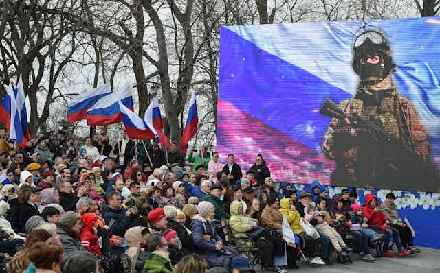 A crownd of people watch a Rusian soldier on a video screen.