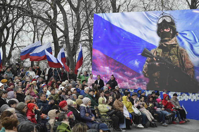 A crownd of people watch a Rusian soldier on a video screen.