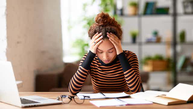 A young woman sitting at a table with papers and a laptop, holding her head in her hands.
