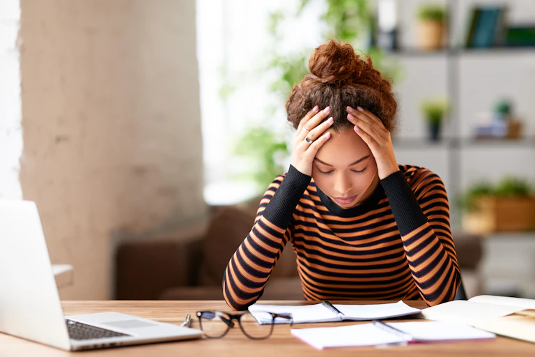 A young woman sitting at a table with papers and a laptop, holding her head in her hands.