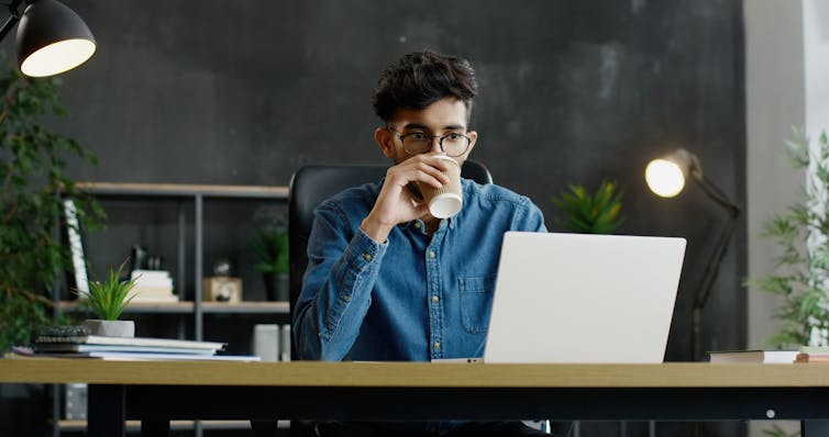 A young man sits at a desk drinking a coffee.