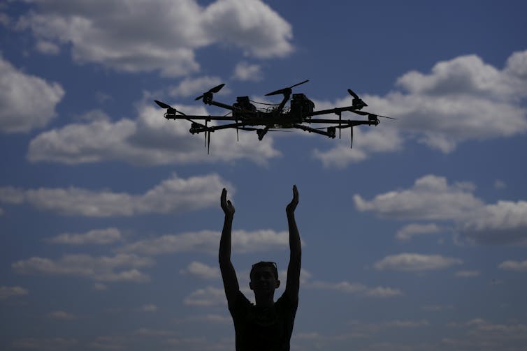 A young man stands with out-stretched arms with a drone in the sky in front of him.