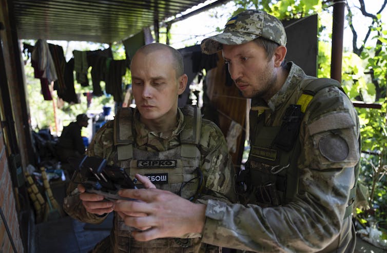 Two soldiers dressed in battle fatigues look at a screen in an outdoor shelter.