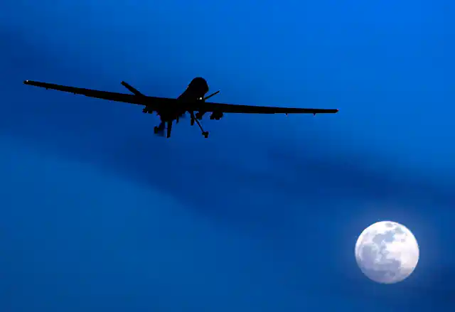 A drone is seen in silhouette in the sky with a full moon beneath it.