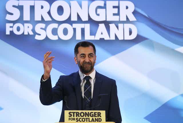 Hamza Yousaf waving in front of a sign that reads 'stronger for Scotland' 